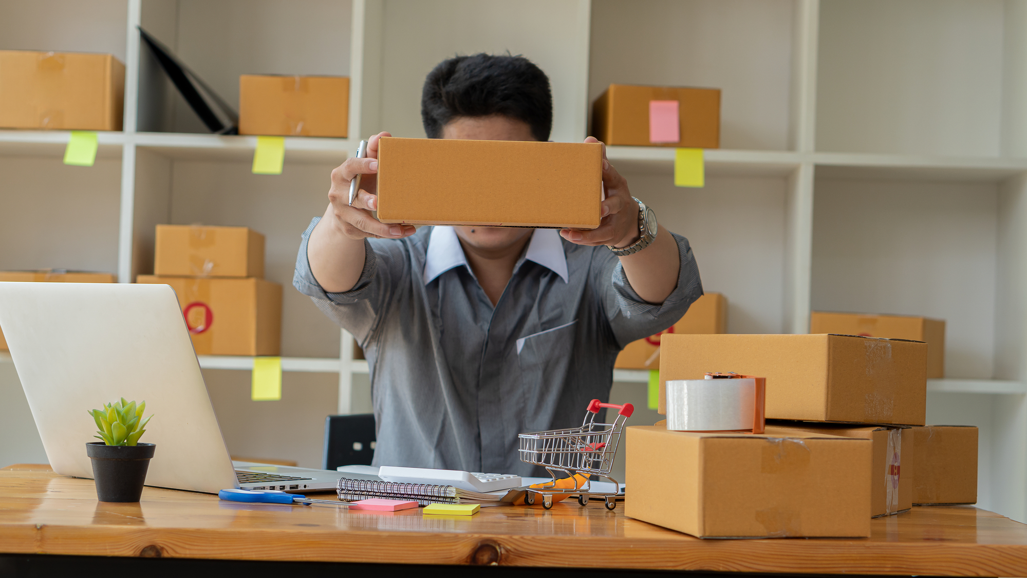 Man holding a box above a desk containing a laptop and mini shopping cart.