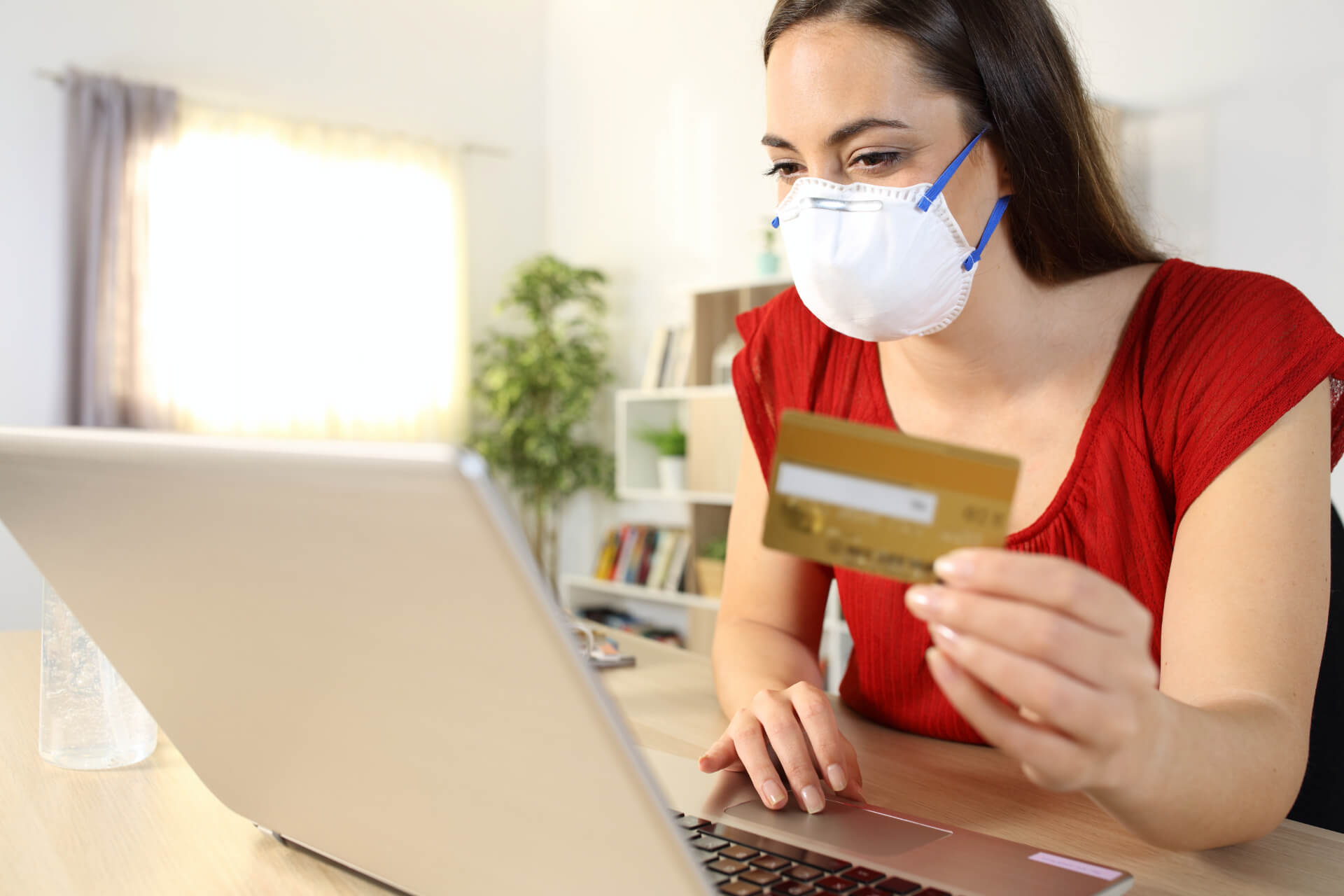 Woman paying online with a card on laptop wearing a mask in covid-19 confinement sitting on a desk at home